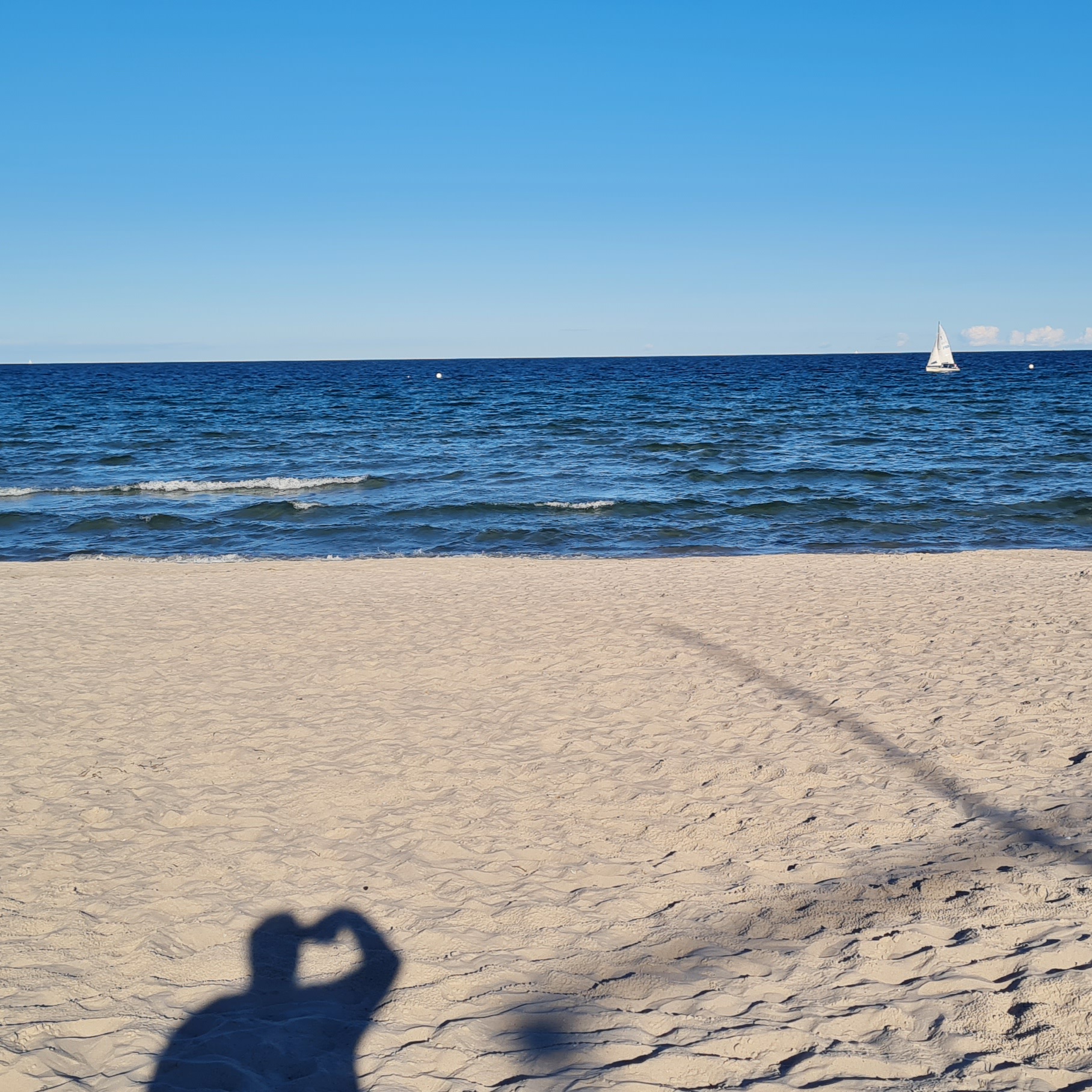 Wellen auf der Ostsee, ein Segelboot am Horizont, blauer Himmel und der Schatten des Fotografs fällt auf den Sandstrand.