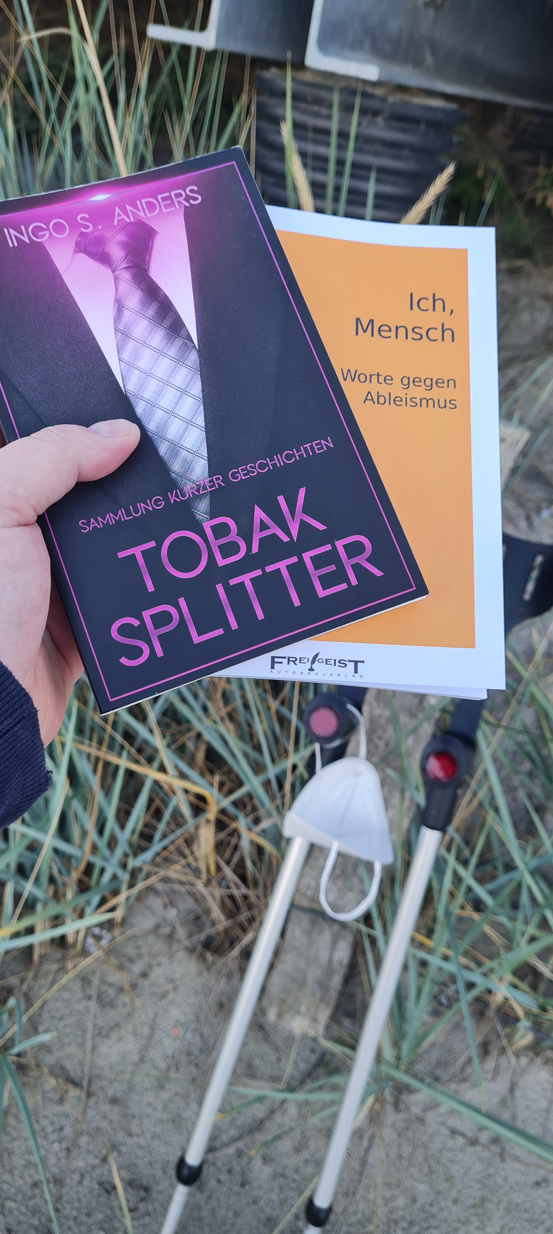 Zwei Bücher in einer Hand, Tobaksplitter von Ingo S. Anders und Ich, Mensch Worte gegen Ableismus, erschienen beim Frei!Geist Autorenverlag. Im Hintergrund lehnen meine Krücken an einem Dünengras-Schutzzaun, an einer hängt eine FFP2-Maske.