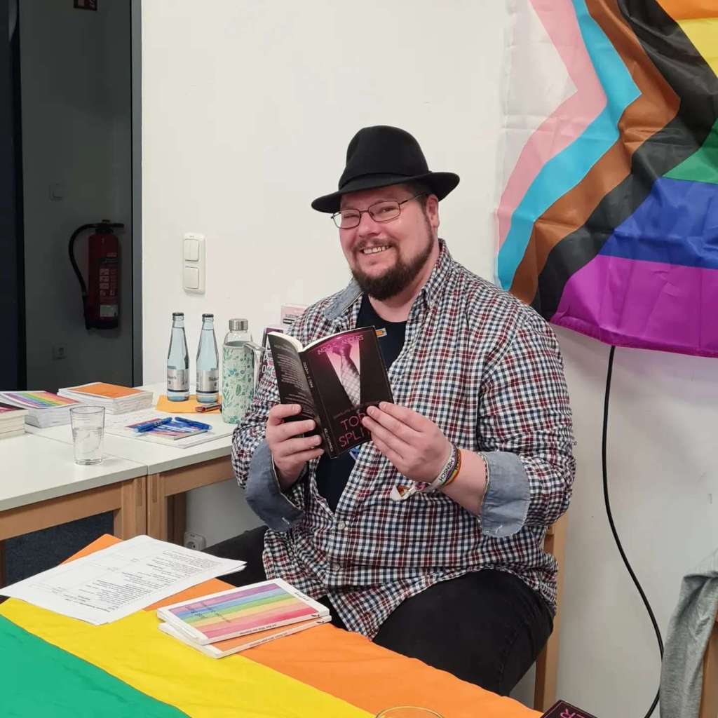 Ein Mann mit Hut hält ein Buch in den Händen. Er lacht in die Kamera.