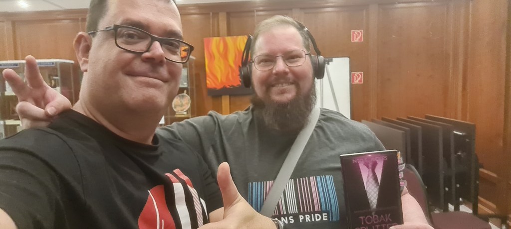 Zwei Männer lächeln in die Kamera. Einer glattrasiert mit Brille, einer mit Bart, Brille und Kopfhörern. Der bärtige Mann trägt ein trans pride-T-Shirt und hält das Buch Tobaksplitter in der Hand. Der rasierte Mann reckt den Daumen nach oben.