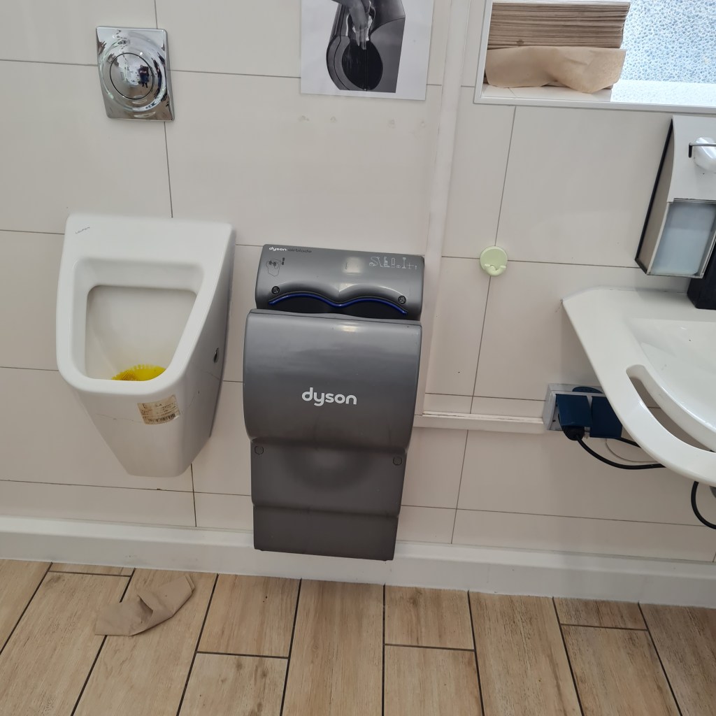 Ein Handtrockner neben einem Urinal auf gleicher Höhe.