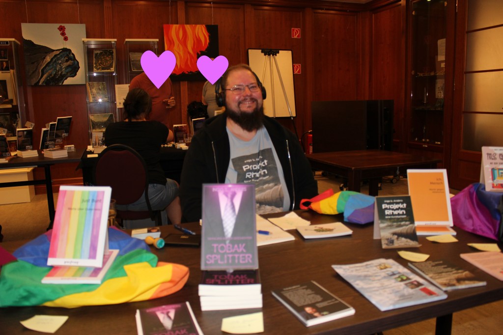 Ein bärtiger Mann mit Brille und Kopfhörern sitzt hinter seinem Büchertisch. Er trägt ein T-Shirt mit aufgedrucktem Buchcover.