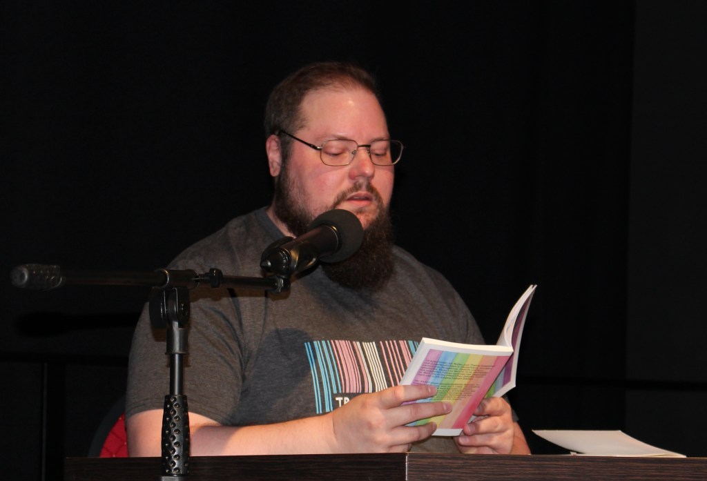 Ingo S. Anders mit Bart und Brille liest aus einem in Regenbogenfarben gestreiften Buch. Er trägt ein T-Shirt mit einem Barcode in den trans Farben.