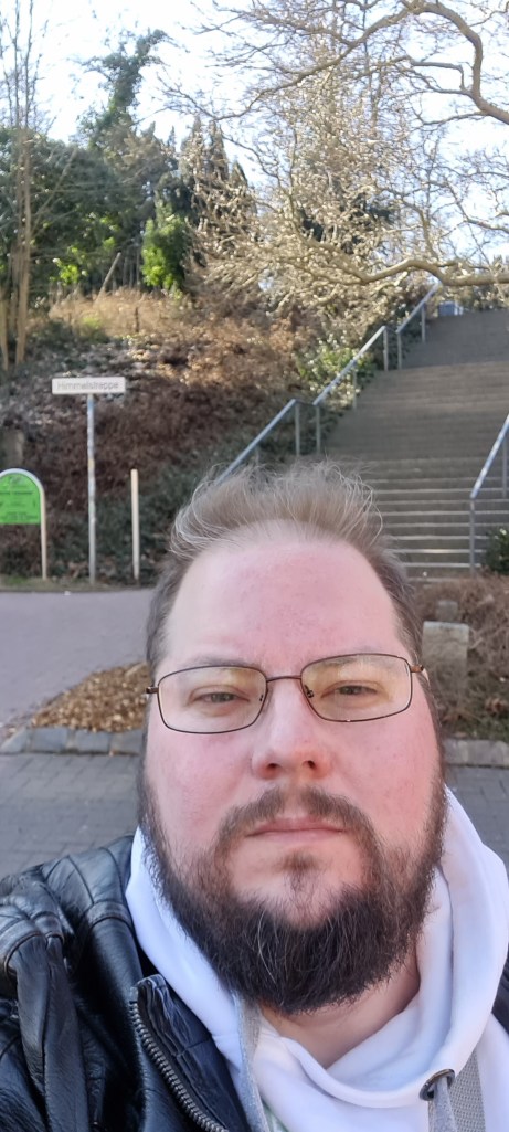 Selfie eines Mannes im Frühling, im Hintergrund eine lange, steile Treppe. Auf einem Schild am Beginn steht: Himmelstreppe