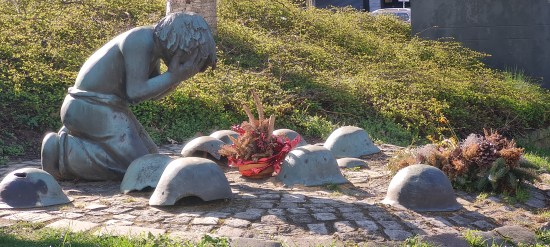 Eine Bronze: Seitlich zu sehen ein knieendes Kind, das Gesicht in den Händen verborgen. Um es herum liegen beschädigte Helme gefallener Soldaten. Zwei Gestecke wurden abgelegt.