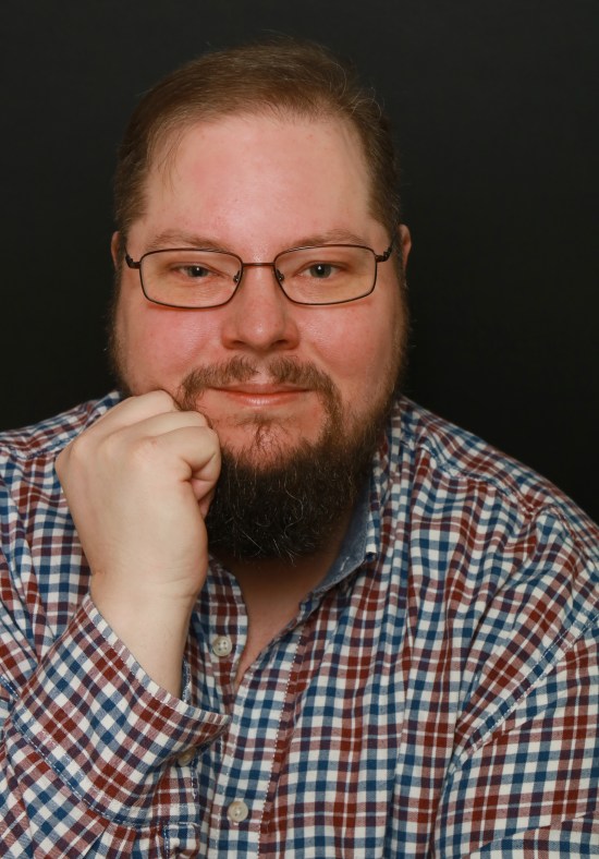 Mann mit heller Haut im kariertem Hemd vor schwarzem Hintergrund. Er trägt eine Brille und einen getrimmten Bart. Seine Haare sind dunkelblond.